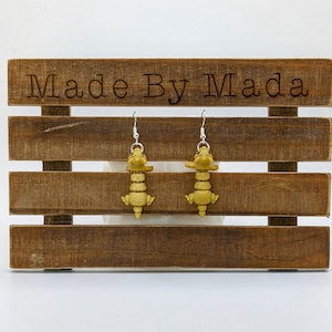 Könnte beinhalten: Ein Paar goldene Alligator-Ohrringe, die an einem Holzständer mit dem Text "Made By Mada" hängen.