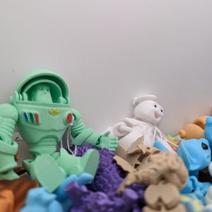 May include: A green astronaut toy with a white helmet and a red, blue, and green stripe on the chest. The toy is surrounded by a variety of other colorful toys, including a white ghost-like figure with a red mouth.