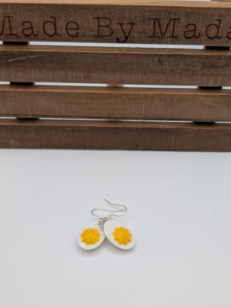 May include: A pair of silver dangle earrings featuring a white ceramic egg with a yellow yolk. The earrings are a fun and quirky accessory for anyone who loves food.