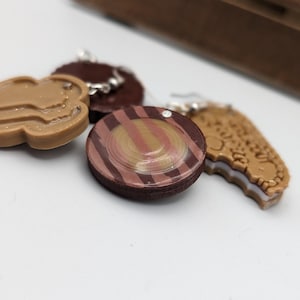 May include: Three pairs of earrings shaped like different cookies. One pair is a brown and white cookie with a swirl design, another pair is a brown cookie with a swirl design, and the last pair is a brown cookie with a white filling.