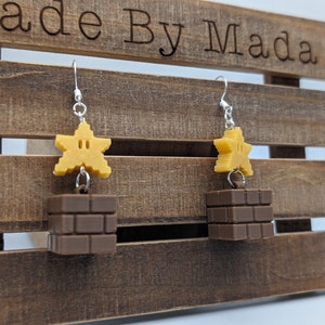 以下が含まれることがあります： 茶色のレンガブロックと顔のある黄色の星がついたイヤリングのペア。イヤリングは木製の背景からぶら下がっています。