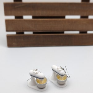May include: A pair of white stand mixer earrings with silver hooks. The mixer bowls are filled with yellow batter.