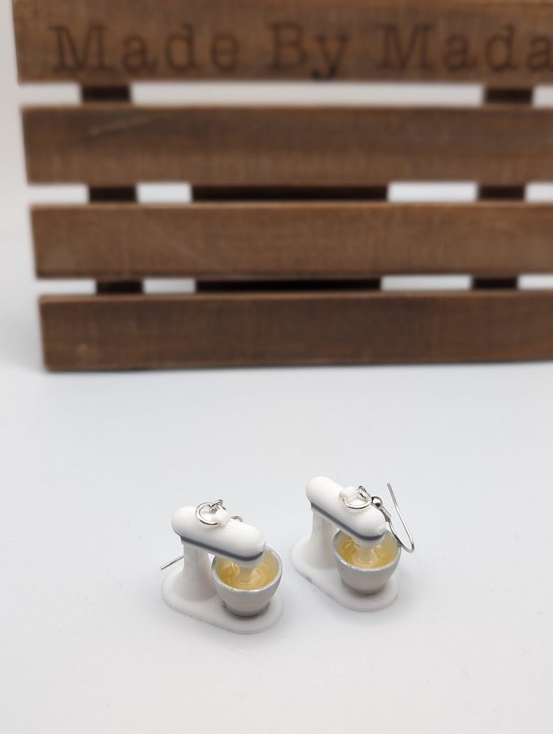 May include: A pair of white stand mixer earrings with silver hooks. The mixer bowls are filled with yellow batter.