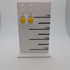 May include: A pair of silver earrings with white and yellow round charms. The charms are shaped like a fried egg with a yellow yolk. The earrings are displayed on a white card with a ruler marked in inches.