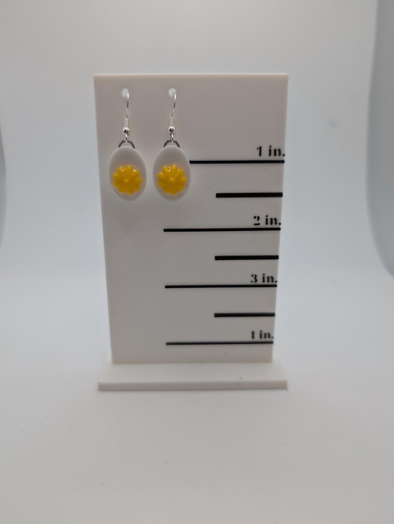 May include: A pair of silver earrings with white and yellow round charms. The charms are shaped like a fried egg with a yellow yolk. The earrings are displayed on a white card with a ruler marked in inches.