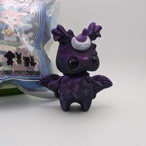 May include: A small, purple, 3D printed creature with a crescent moon on its head and large, black eyes. The creature has wings and is standing on a white surface.