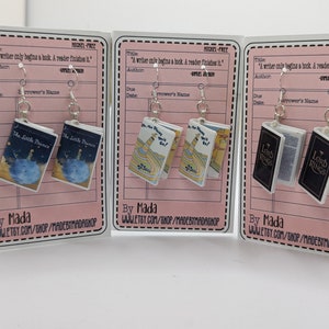 May include: Three pairs of book-themed earrings on pink card stock with a white border. The earrings are nickel-free and feature miniature versions of "The Little Prince", "Oh, the Places You'll Go!" and "The Lord of the Rings" books. Each card has the text "A writer only begins a book. A reader finishes it." and "By Mada" with a website address.