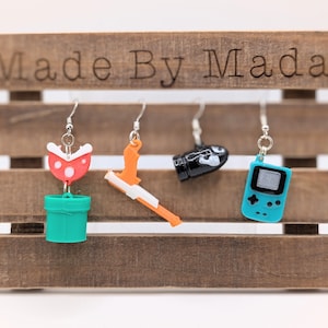 May include: Four pairs of earrings featuring video game characters. The earrings are displayed on a wooden shelf with the text "Made By Mada". The earrings feature a green pipe with a red flower, a black and white gun, a black bomb, and a blue Game Boy.