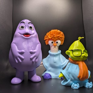 Puede incluir: Tres figuras coloridas de personajes de dibujos animados. La primera es una figura morada en forma de mancha con una cara sonriente. La segunda es un personaje pelirrojo con gafas y una túnica azul. La tercera es un personaje verde con gafas y una bufanda naranja.