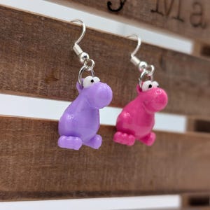 Può includere: Un paio di orecchini pendenti con un ciondolo a forma di ippopotamo viola e rosa. Gli orecchini sono in argento e hanno una chiusura a gancio.