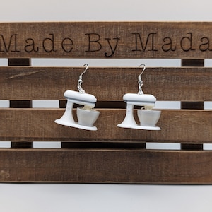 May include: A pair of white and gray stand mixer earrings. The earrings are hanging from a wooden background with the text "Made By Mada" on the top.