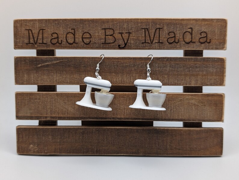 May include: A pair of white and gray stand mixer earrings. The earrings are hanging from a wooden background with the text "Made By Mada" on the top.