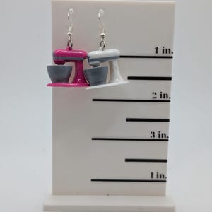 May include: A pair of dangle earrings featuring miniature kitchen stand mixers. One mixer is pink with a gray bowl, and the other is white with a gray bowl. The earrings are displayed on a white ruler with markings in inches.