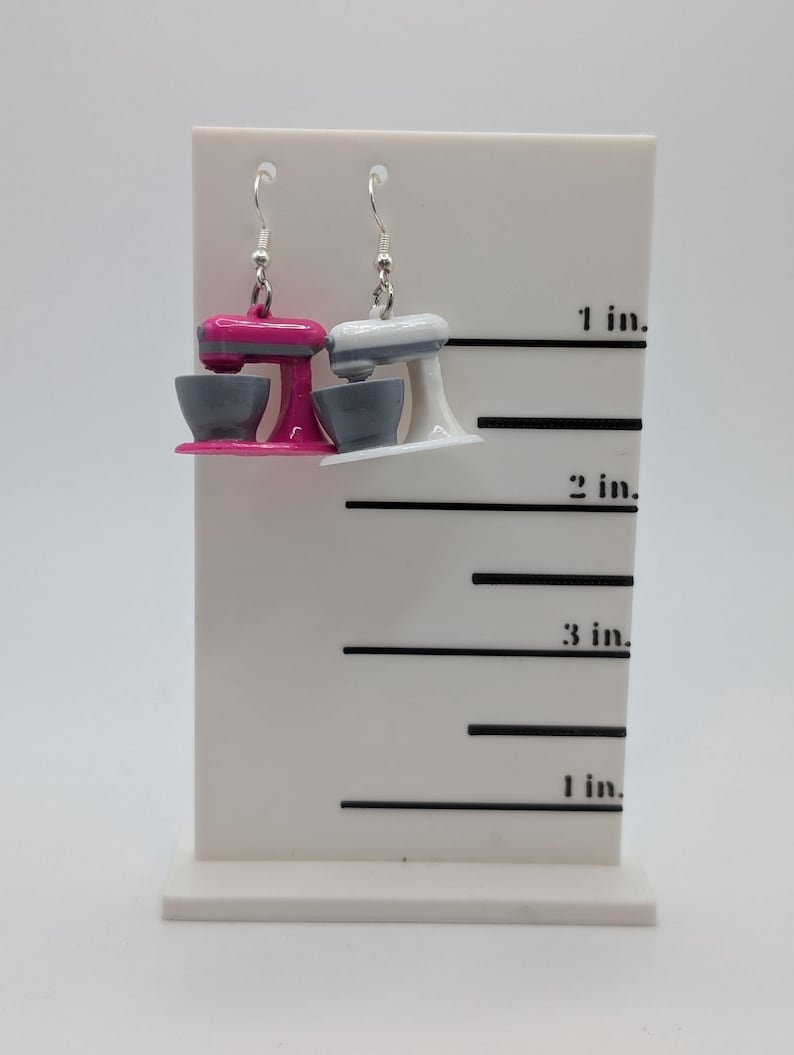 May include: A pair of dangle earrings featuring miniature kitchen stand mixers. One mixer is pink with a gray bowl, and the other is white with a gray bowl. The earrings are displayed on a white ruler with markings in inches.