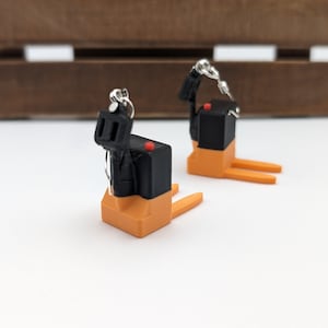 Puede incluir: Un par de pendientes en forma de carretilla elevadora en miniatura con cuerpo negro, horquillas naranjas y un pequeño botón rojo. Cada pendiente está sujeto a un gancho de metal plateado, diseñado para colgar verticalmente.