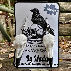 Könnte beinhalten: Ein Paar schwarze und weiße Rabenschädel-Ohrringe auf einer weißen Karte mit einer schwarzen Rabenillustration und dem Text "Nickel Free" und "By Madebymaa".