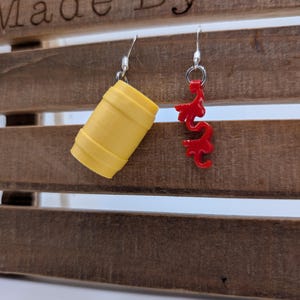 Puede incluir: Un par de pendientes con un barril amarillo y un dije de dragón rojo. Los pendientes están colgados en un estante de madera con el texto "Made By M" grabado en la parte superior.