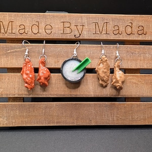 May include: A wooden display stand with the text "Made By Mada" engraved on the top. The stand holds five pairs of earrings: two red chicken wing earrings, a small bowl earring with white and green details, and two brown chicken wing earrings.