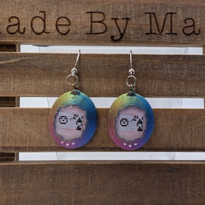 May include: A pair of iridescent rainbow earrings with a pink and white design. The earrings are shaped like a Tamagotchi, a popular virtual pet toy from the 1990s.