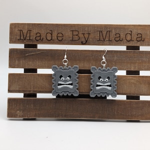 May include: A pair of gray 3D printed earrings shaped like a square with a frowning face. The earrings are hanging on a wooden display stand with the text "Made By Mada" engraved on the top.