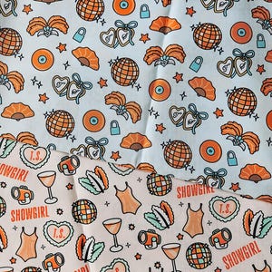 May include: Two patterned fabrics, one light blue and one light pink, feature a repeating design of orange disco balls, fans, hearts, cameras, and the word "SHOWGIRL". The light blue fabric has a similar pattern.