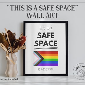 May include: Black framed wall art print with the text "This is a Safe Space" and a rainbow pride flag with a transgender pride flag incorporated into the design. The text "Be uniquely you!" is below the flag. The print is on a white background and has a black frame. The print is on a white wall with a vase of dried flowers in front of it.