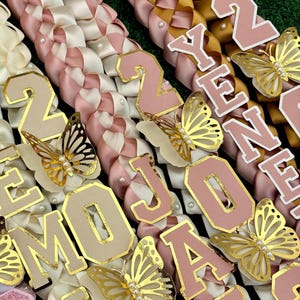 May include: Close-up of several graduation leis with woven ribbons in cream, pink, and gold. Each lei features gold-trimmed letters and butterfly accents. Some leis spell out "2024" and "JOANNA".