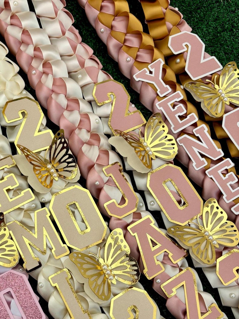 May include: Close-up of several graduation leis with woven ribbons in cream, pink, and gold. Each lei features gold-trimmed letters and butterfly accents. Some leis spell out "2024" and "JOANNA".
