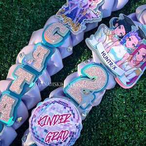 May include: A close-up of two graduation leis with light purple ribbons. The leis feature glittery letters and numbers, along with cartoon graphics and the words "Kinder Grad". The leis are decorated with rhinestones.