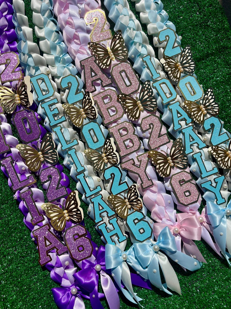May include: A collection of colorful ribbon leis with personalized names and numbers. Each lei features glittery letters and numbers, butterfly accents, and decorative bows. The leis are in shades of purple, blue, and pink, perfect for graduation or special events.
