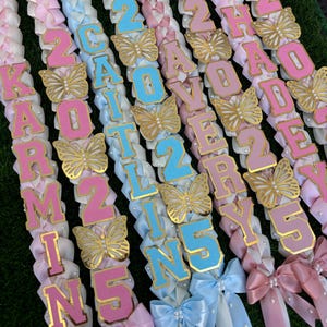 May include: Several leis with pink, white, and blue ribbons are displayed. Each lei features a name and the number 25, with gold butterfly accents. The names are spelled out in pink and blue letters.