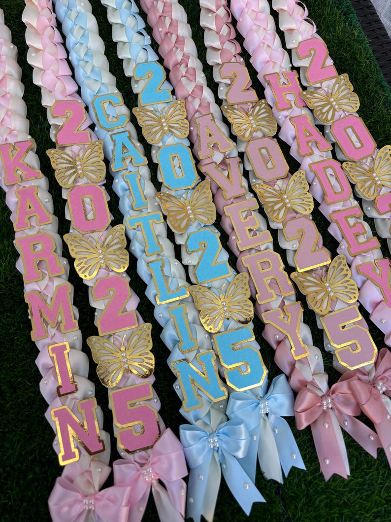 May include: Several leis with pink, white, and blue ribbons are displayed. Each lei features a name and the number 25, with gold butterfly accents. The names are spelled out in pink and blue letters.