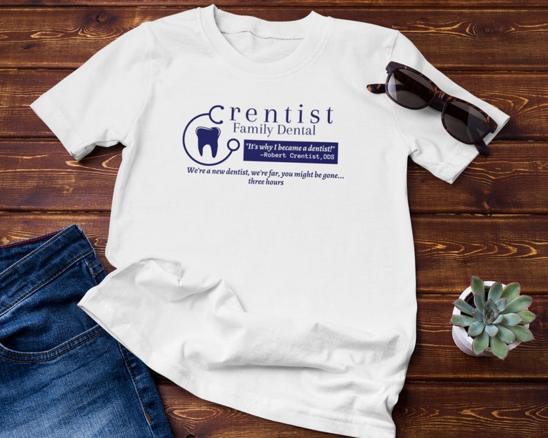 May include: White t-shirt with a blue graphic of a dentist's logo and the text "Crentist Family Dental" and "It's why I became a dentist! -Robert Crentist, DDS We're a new dentist, we're far, you might be gone... three hours"