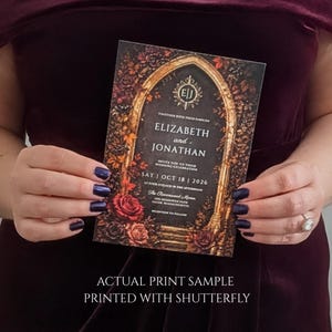 May include: A rectangular wedding invitation card featuring a dark gothic archway design surrounded by deep red and orange autumn roses and foliage. The text reads: Elizabeth and Jonathan, Sat Oct 18 2026, The Ravenswood Manor, Salem, Massachusetts.