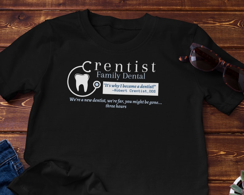 May include: Black t-shirt with a white graphic of a tooth and a stethoscope. The text "Crentist Family Dental" is written in white above the graphic. The text "It's why I became a dentist! -Robert Crentist, DDS" is written in blue below the graphic. The text "We're a new dentist, we're far, you might be gone... three hours" is written in white below the blue text.
