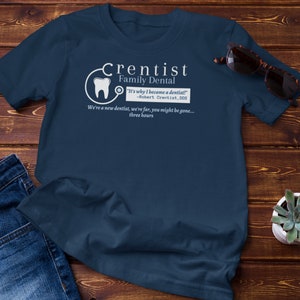 May include: A navy blue t-shirt with a white graphic of a tooth and stethoscope. The text "Crentist Family Dental" is written in white above the graphic. The text "It's why I became a dentist!" is written in white below the graphic. The text "Robert Crentist, DDS" is written in white below the quote. The text "We're a new dentist, we're far, you might be gone... three hours" is written in white below the DDS text.