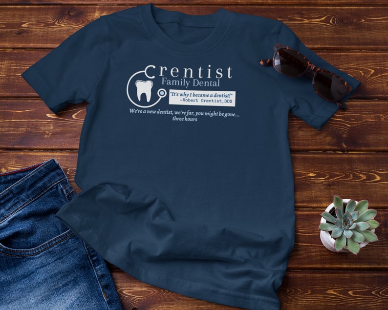 May include: A navy blue t-shirt with a white graphic of a tooth and stethoscope. The text "Crentist Family Dental" is written in white above the graphic. The text "It's why I became a dentist!" is written in white below the graphic. The text "Robert Crentist, DDS" is written in white below the quote. The text "We're a new dentist, we're far, you might be gone... three hours" is written in white below the DDS text.