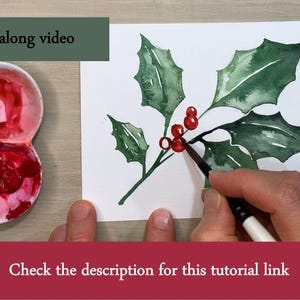 Pode incluir: Pintura em aquarela de azevinho com folhas verdes e bagas vermelhas. Uma m&atilde;o est&aacute; pintando as bagas com um pincel. O texto na imagem diz "Paint along video" e "Check the description for this tutorial link".
