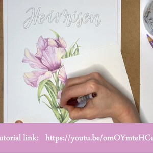 May include: A hand is colouring a drawing of pink tulips with green leaves. The drawing is on a white sheet of paper with the words "He is risen" written in cursive at the top. The tutorial link is "https://youtu.be/omOYmteHCeo".