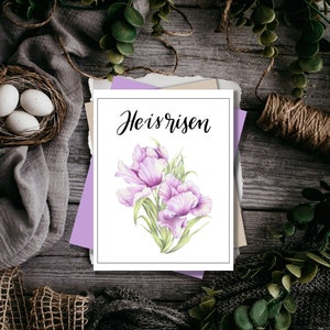 May include: A white card with the text "He is risen" in black calligraphy. The card is decorated with a watercolour illustration of pink flowers.