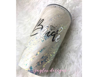 May include: A white tumbler with a glittery finish and a black and silver vinyl design that says "Bride" with a diamond symbol.