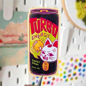 Peut inclure: Un autocollant coloré représentant une canette de boisson énergisante. La canette est noire avec les mots "TURBO ENERGY" en rouge et jaune. Un chat blanc avec des détails rouges est sur la canette, et le texte "ENERGY DRINK 12 FL. OZ." est également présent.