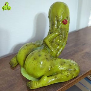May include: A whimsical, green pickle figurine with a suggestive pose, featuring red lips. The sculpture is detailed with a textured surface and sits on a green base. The logo "CRLES" is visible in the upper left corner.
