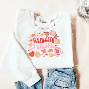 Op de afbeelding: Witte sweatshirt met de tekst "Cafecito y Chisme" in rood en roze, omringd door illustraties van gebak, harten en rozen. Een zilveren ketting en een blauwe jeans zijn ook zichtbaar.