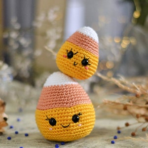 May include: Two crocheted candy corn toys with smiling faces. The larger candy corn is orange, yellow, and white. The smaller candy corn is stacked on top of the larger one. The candy corn toys are on a blue and white surface.
