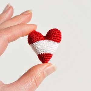 May include: A small crocheted heart in red and white, resembling the Austrian flag, is held between two fingers.