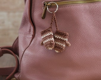 Puede incluir: Una mochila de cuero rosa con un pequeño llavero de manoplas de ganchillo. Las manoplas son marrones y blancas con un patrón a rayas.