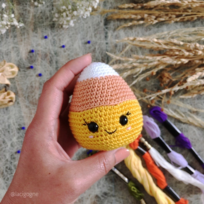 May include: A crocheted candy corn toy with a white top, orange middle, and yellow bottom. The toy has a smiling face with black eyes and a black mouth.