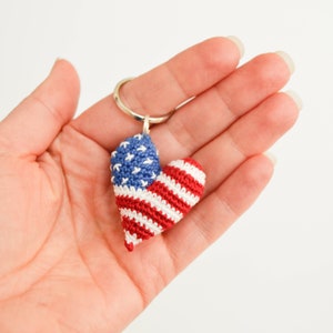 May include: A crocheted heart-shaped keychain with the American flag design. The keychain is red, white, and blue and is attached to a silver key ring.