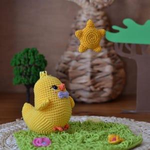 Puede incluir: Un pollito amarillo de ganchillo hecho a mano con una pajarita morada se sienta sobre una base verde de ganchillo con una flor y una estrella amarilla. Un conejo tejido y un peque&ntilde;o &aacute;rbol est&aacute;n en el fondo. Decoraci&oacute;n caprichosa.